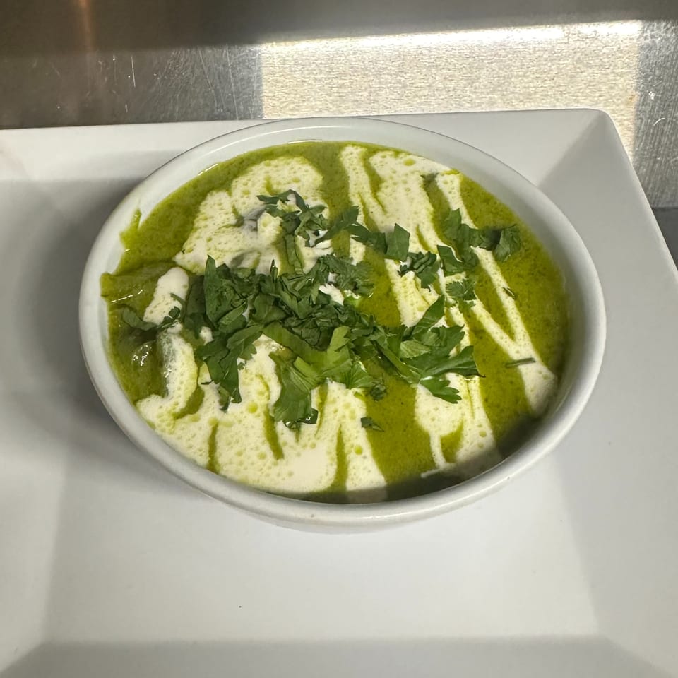 Green soup with white cream swirls and fresh parsley in a white bowl on a white plate.