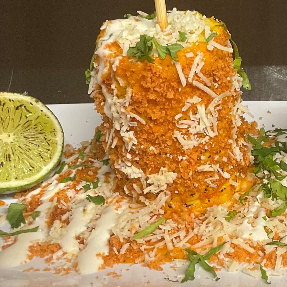 Mexican street corn (elote) with orange seasoning, cheese, white sauce, and herbs, served with a lime.