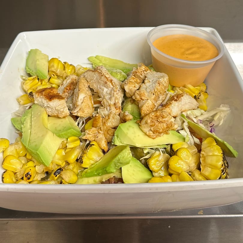 Grilled chicken, charred corn, and avocado slices in a white bowl with a side of orange dressing.